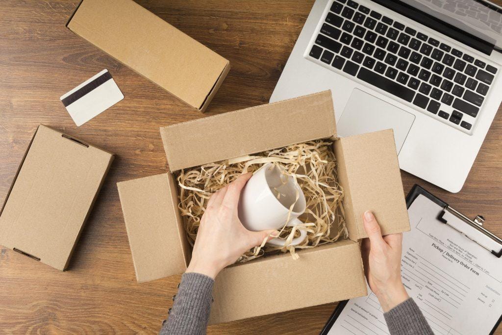 Mujer desempaquetando un producto en caja con laptop y formulario de envío.